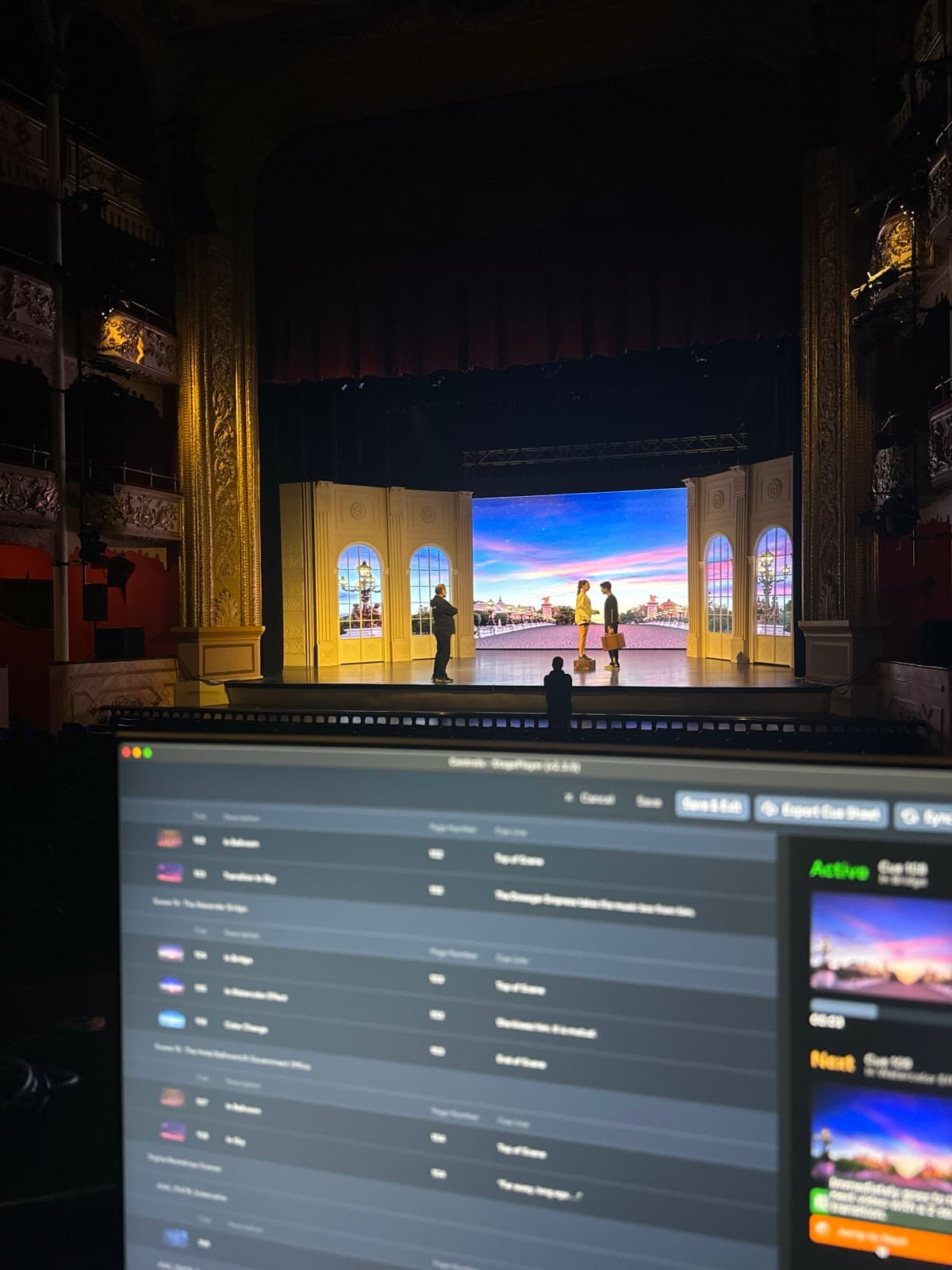 A technician's laptop running a StagePlayer cue list in a theatre booth, with digital scenery projected on the stage beyond